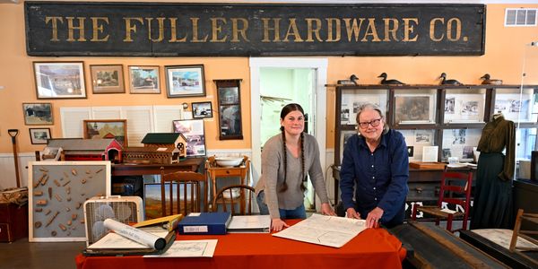 Two women stand behind a table with blueprints in a vintage hardware store.