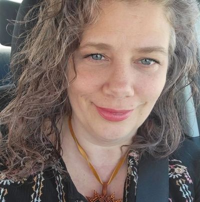 Woman with curly hair smiling in a car, wearing a black patterned top and sun-shaped pendant.