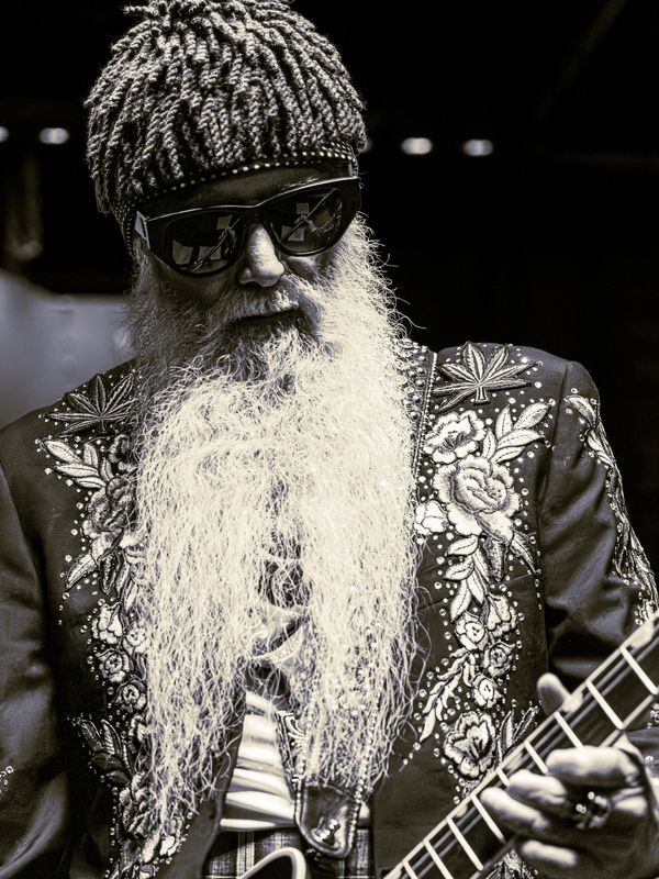 Bearded musician wearing sunglasses and intricate jacket playing guitar.