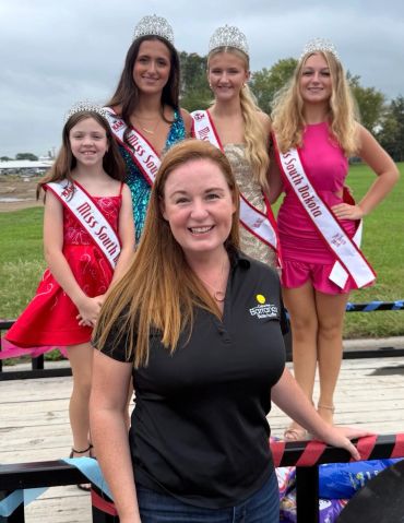 Four beauty queens and a woman smiling outdoors.