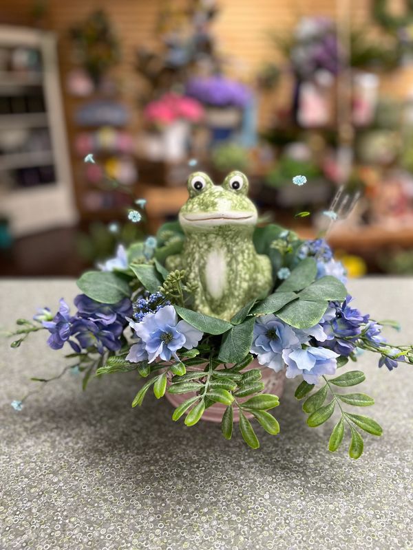 Ceramic frog figurine surrounded by blue flowers and green leaves in a decorative arrangement.