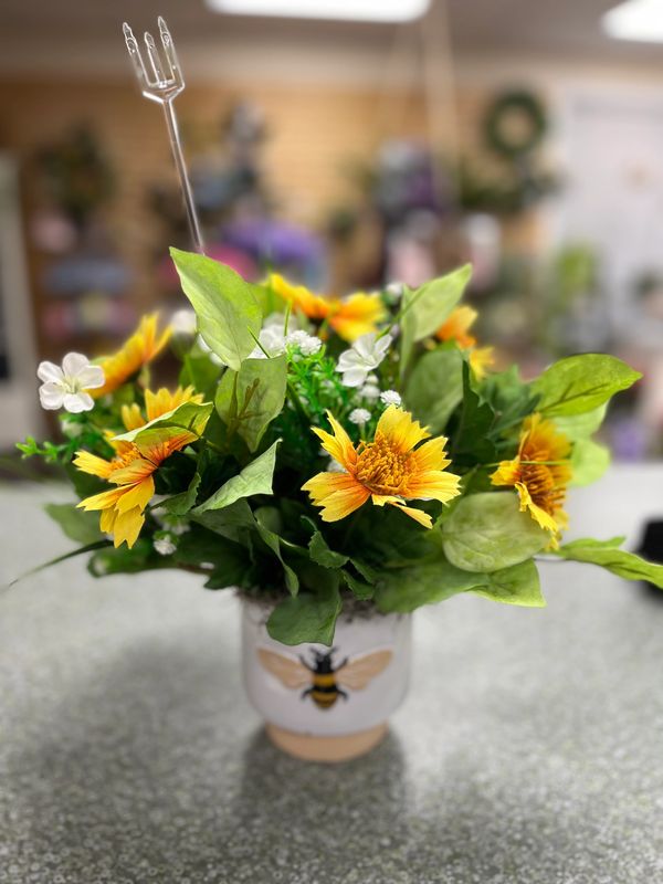 Bright yellow flowers in a bee-themed pot with a small pitchfork decoration.