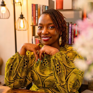 Smiling woman in a vibrant patterned blouse sits at a desk with books and a laptop.