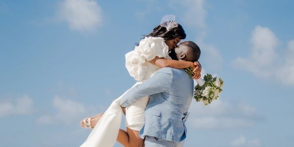 Groom lifts bride joyfully against a blue sky on their wedding day.