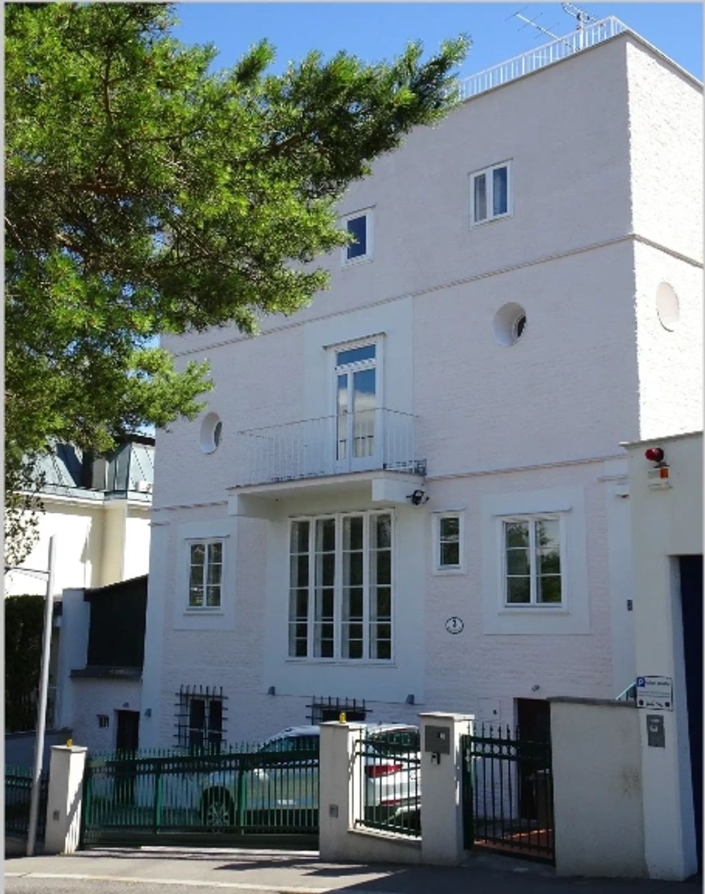White multi-story house with green gate and parked car under bright daylight.
