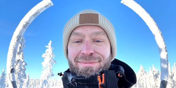 Man in winter clothing smiling outdoors with snowy trees and clear blue sky.