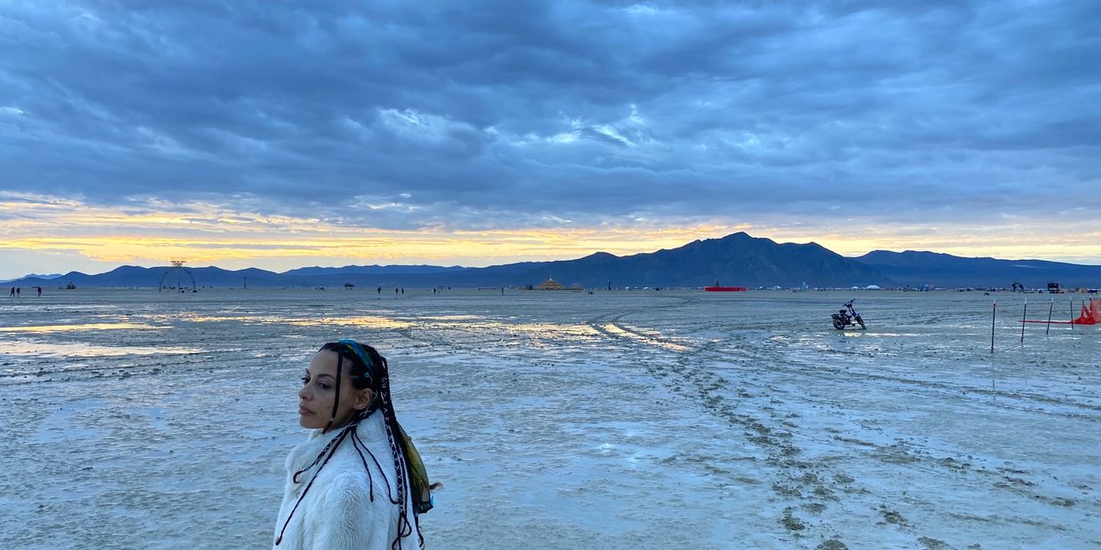 Woman in white coat stands on vast desert playa at sunset with mountains in background.