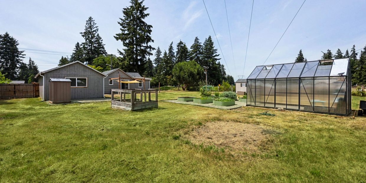 Backyard with a greenhouse, raised garden beds, and a small shed near houses under power lines.
