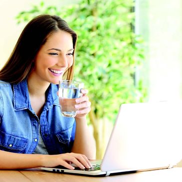 Young woman smiling while using laptop and holding a glass of water.