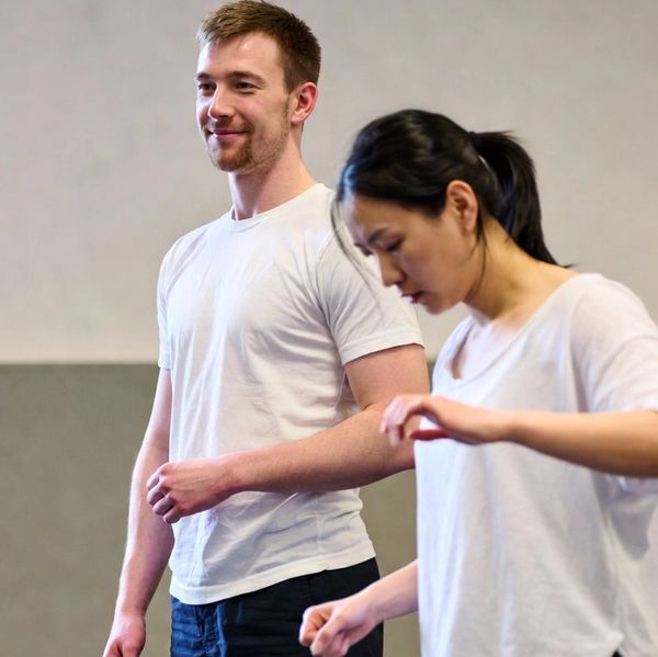 Two people practicing movement or dance indoors, wearing casual white tops and dark pants.