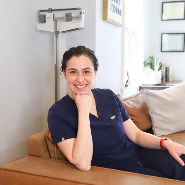 Smiling healthcare professional in navy scrubs sitting on a couch.