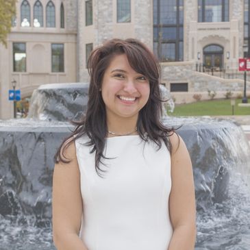 A smiling woman in a white dress stands in front of a fountain and building.