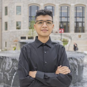 Young man in a black shirt stands confidently with arms crossed outdoors.