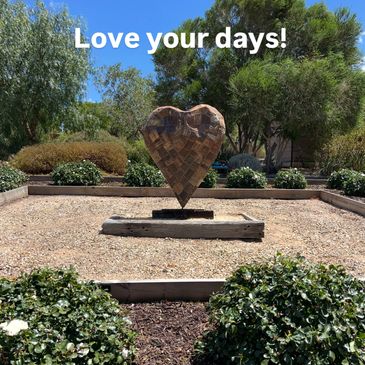 Heart-shaped sculpture in a sunny garden with 'Love your days!' text.