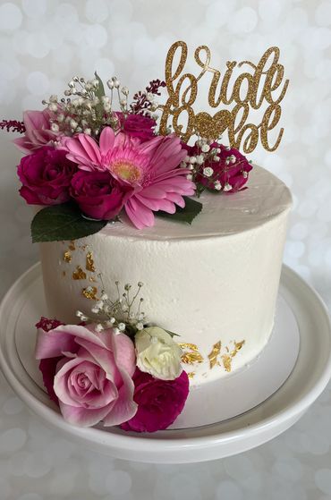 A white buttercream cake with gold leaf detail, pink flowers and an acrylic topper. 