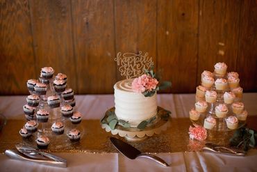 Simple white buttercream cake with fresh flowers and acrylic topper surrounded by mini pink cupcakes
