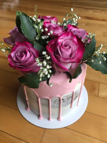 A white semi naked buttercream cake with fresh pink flowers and pink drip. 