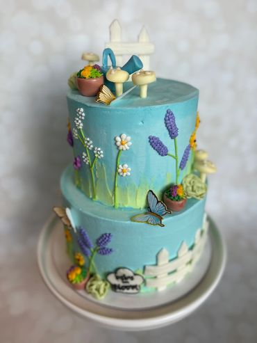 Two-tier garden-themed cake with flowers, mushrooms, and a white picket fence from fondant. 