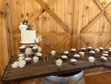 A white buttercream cake surrounded by cupcakes in a rustic venue. 