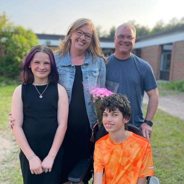 ID: Photo is of Karin and her husband and two children.  They are standing in the sun in front of a 