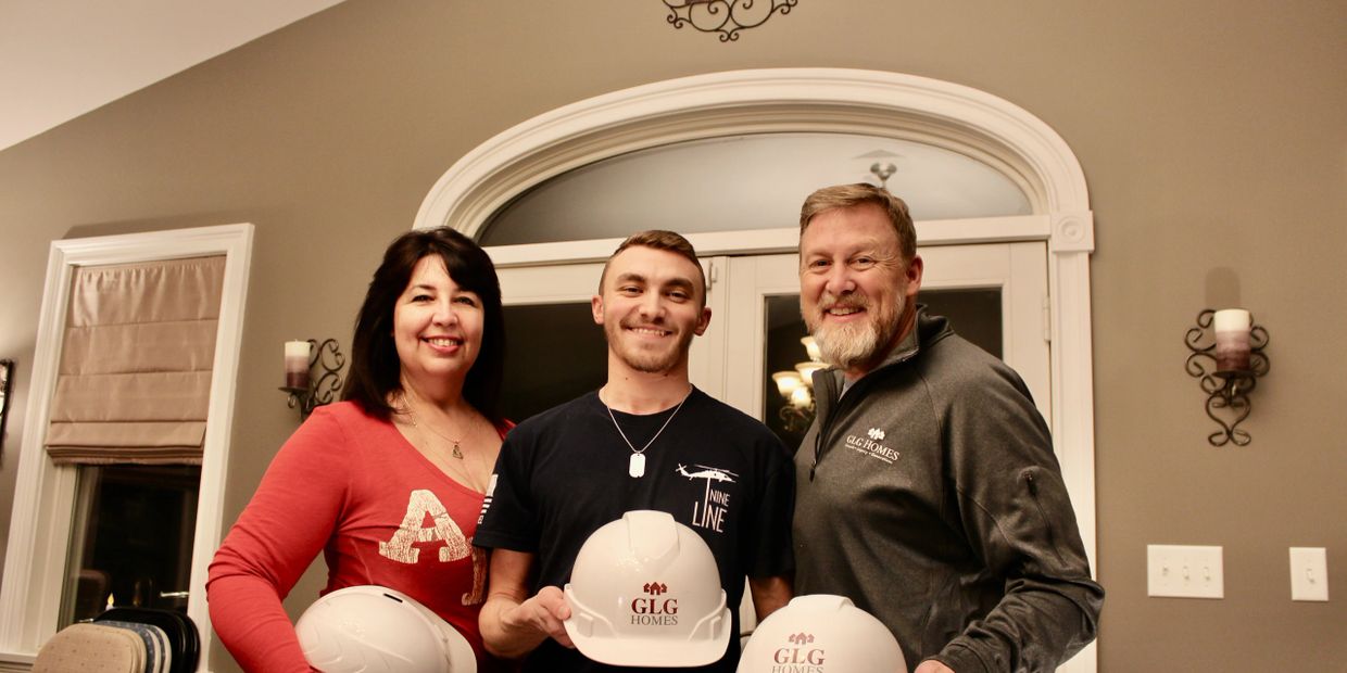 Three people smiling, holding GLG Homes hard hats indoors.