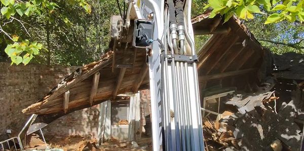 Excavator demolishing a wooden roof in a sunny, leafy area.