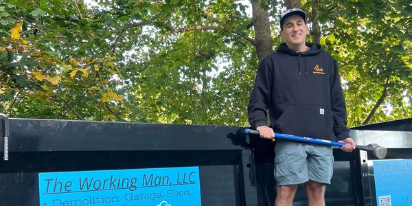 Man in a black hoodie holding a tool, standing by a dumpster with a business sign.