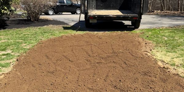 Freshly tilled soil patch next to a driveway with a trailer and a parked vehicle.