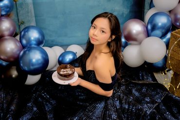 A woman in a black dress holding a birthday cake surrounded by balloons.