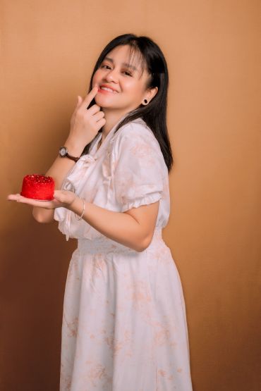 Young woman in white dress holding a small red cake, smiling.