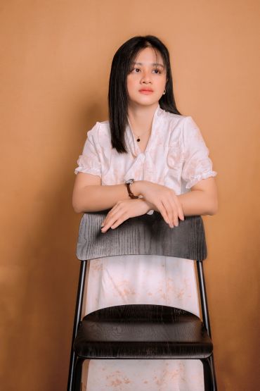 Young woman in white dress leaning on chair against brown background.