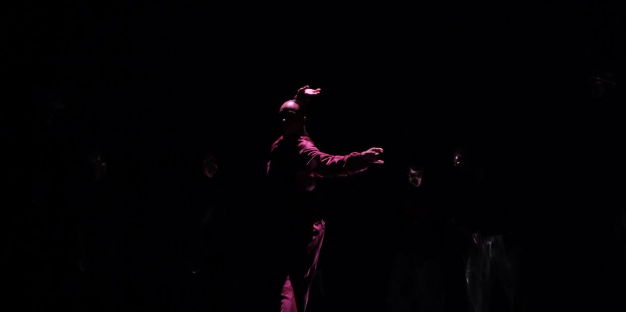 A dancer illuminated in purple light, surrounded by darkness and faint figures.