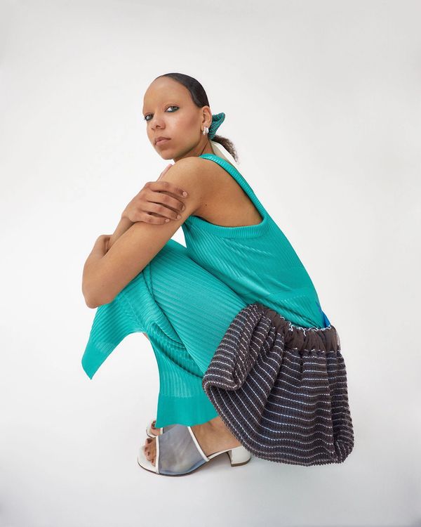 Woman in turquoise dress crouching with a textured bag and white heeled sandals.