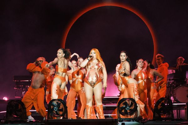 Singer and dancers perform on stage with vibrant orange costumes and a glowing circular backdrop.