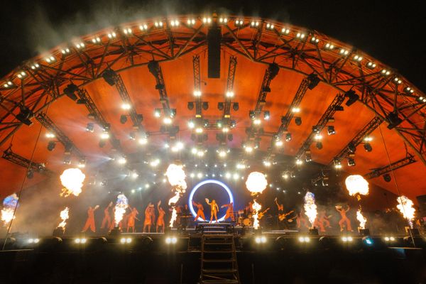 Performers on a brightly lit stage with flames and an orange canopy overhead.