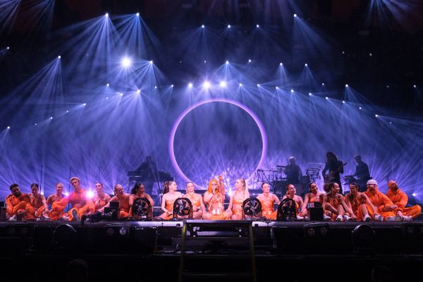 A group of performers in orange outfits on stage with dramatic blue lighting and a circular backdrop.