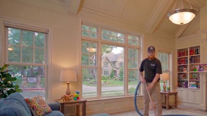 A man vacuuming a cozy living room with large windows overlooking a suburban neighborhood.