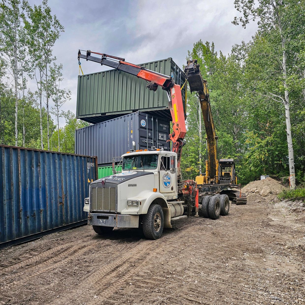 Truck with crane lifting a shipping container at a forested construction site.