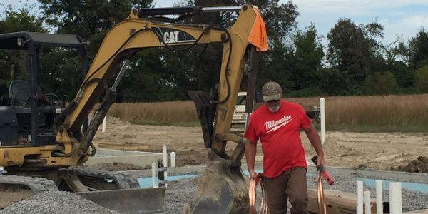 Construction worker carrying copper tubing near excavator on gravel site.