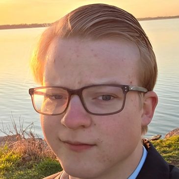 Young person with glasses near a calm lake during sunset.