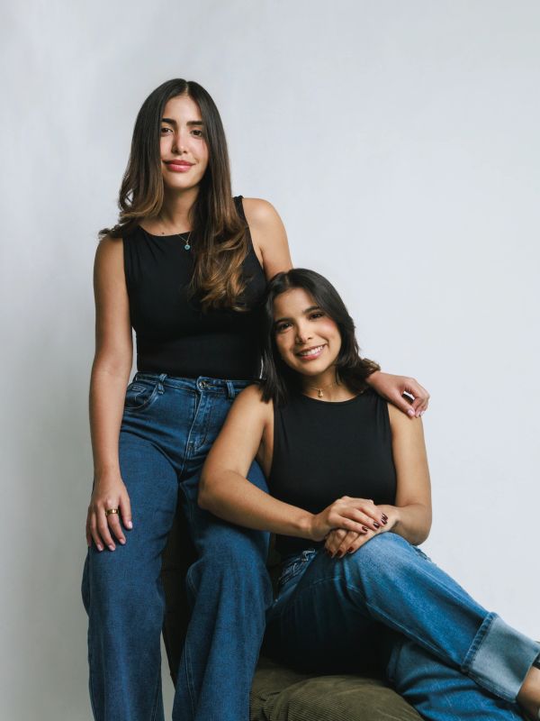 Two women in black tops and blue jeans pose against a plain background.
