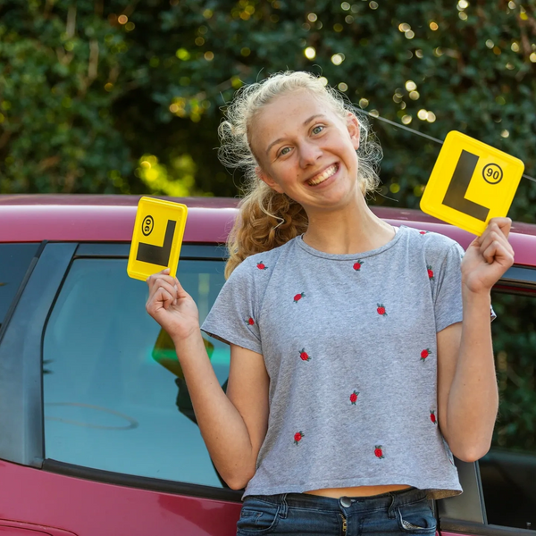 Young woman proudly holding two yellow learner driver plates by a red car.
