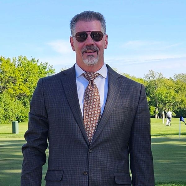 Man in a suit and sunglasses standing outdoors on a golf course.