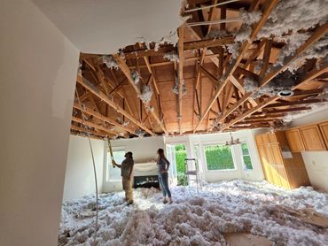 Two people inspect a partially demolished ceiling with insulation scattered on the floor.