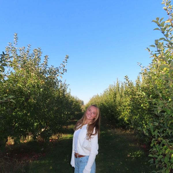 A smiling woman stands in an orchard under a clear blue sky.