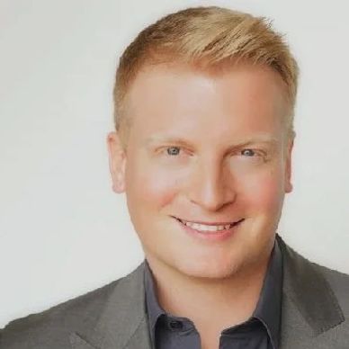 Smiling man with blond hair in a gray blazer and dark shirt.