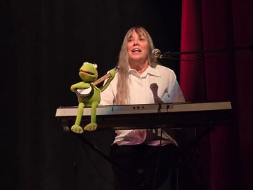 Woman playing keyboard with Kermit the Frog puppet on stage.