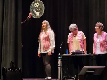 Three women in pink striped vests on stage with a smiley face prop.