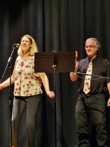 Two people smiling on stage with microphones and music stand.