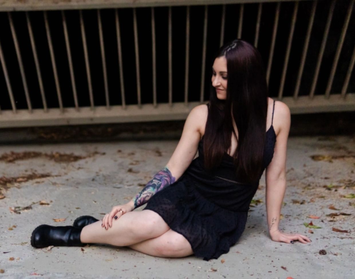 Woman with long dark hair sitting on concrete floor in black dress and boots.